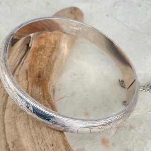 May include: A silver hinged bangle bracelet with an engraved floral pattern. The bracelet is resting on a piece of driftwood. The clasp is visible, along with a safety chain. The bracelet is a classic piece of jewelry.