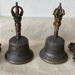 May include: Two ornate bronze bells with detailed carvings and a crown-like handle. Two smaller bronze vajra scepters are also present. The bells have a dark, aged appearance, suggesting a vintage or antique origin. The overall theme is spiritual or religious.