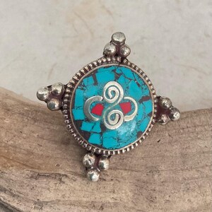 May include: A silver-toned ring with a turquoise stone center. The stone features a silver swirl design and red accents. The ring has a cross-like design with decorative silver details on each end. The ring is set on a wooden surface.