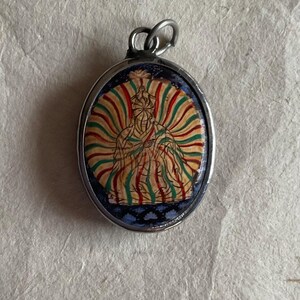 May include: Silver oval pendant with a colorful image of a seated figure with a halo. The figure is surrounded by a radiating pattern of green, red, and yellow stripes.