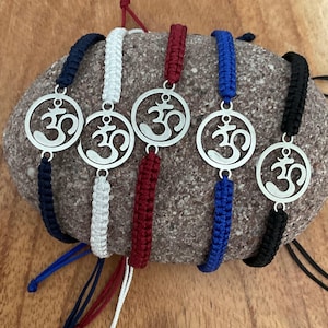 May include: Five braided bracelets with silver Om symbol charms. The bracelets are in navy blue, white, red, blue, and black.