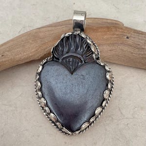 May include: A silver-toned heart pendant with a dark gray center. The pendant has a detailed design around the heart and a loop at the top for a chain. The heart shape is a classic symbol of love and affection.