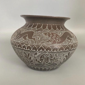 May include: A dark brown ceramic pot featuring intricate white etched designs. The upper band displays running animals, while the lower section depicts a seated figure with an elephant head and multiple arms, surrounded by stylized foliage and leaf patterns.