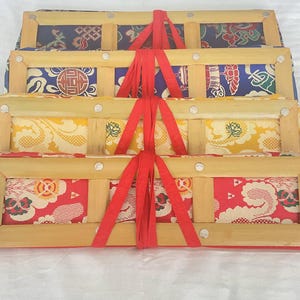 May include: A stack of four rectangular fabric panels, each secured within a light-colored wooden frame. The panels feature vibrant, intricate patterns in red, yellow, blue, and dark blue, all bound together by a bright red fabric ribbon tie.