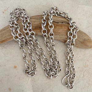 May include: A long silver chain necklace featuring thick, rounded interlocking links. The chain has a polished metallic finish and is draped in a loose, folded arrangement, showcasing the uniform size and circular shape of each individual link.