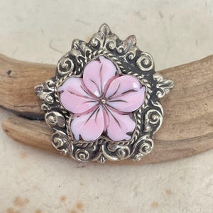 May include: A brooch featuring a central five-petaled light pink flower with carved details, set within an ornate, dark silver-toned metal frame decorated with intricate scrollwork and leaf-like patterns surrounding the floral center.