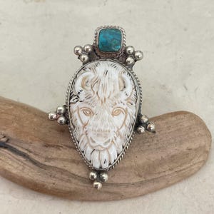 May include: A silver-toned pendant featuring a carved white buffalo head with detailed facial features. A square turquoise stone sits atop the pendant, and small silver beads adorn the sides. The pendant is displayed on a piece of driftwood.