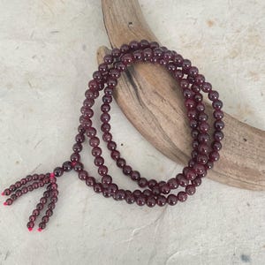 May include: A long strand of deep red gemstone beads, likely garnet, arranged in multiple loops. The beads are round and polished, with a tassel at the end. The necklace rests on a piece of driftwood against a textured, off-white background.