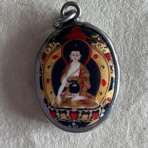 May include: A silver oval pendant with a colorful image of a seated Buddha. The Buddha is wearing a yellow robe and has a black bowl in their lap. The background is blue with gold accents.