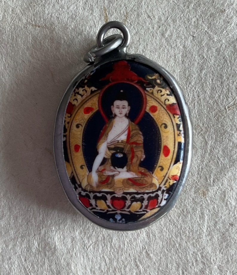 May include: A silver oval pendant with a colorful image of a seated Buddha. The Buddha is wearing a yellow robe and has a black bowl in their lap. The background is blue with gold accents.