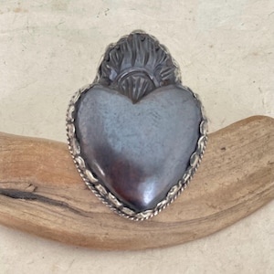 May include: A silver-toned heart-shaped pendant with a dark, iridescent center. The pendant has a detailed, ornate border and a decorative top element. The piece is displayed on a piece of driftwood.