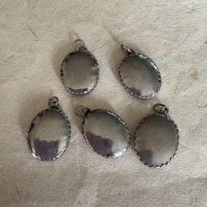 May include: Five silver oval-shaped pendants with a decorative edge. The pendants are blank and have a shiny finish.