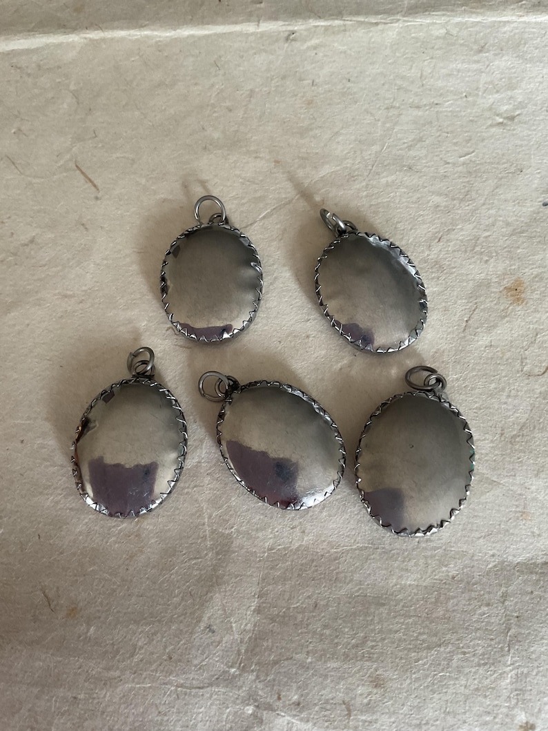 May include: Five silver oval-shaped pendants with a decorative edge. The pendants are blank and have a shiny finish.