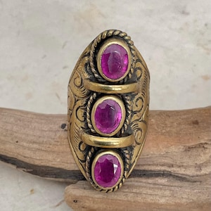 May include: An antique brass ring with three oval, magenta-colored gemstones. The ring has an ornate design with swirling patterns and rope-like detailing. The gemstones are set in a vertical row, adding a touch of elegance to the piece.