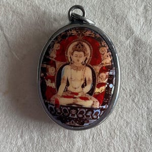 May include: A silver oval pendant with a colorful image of a seated Buddha figure. The Buddha is depicted in a red and gold robe with a halo around their head. The pendant has a detailed border with intricate designs.