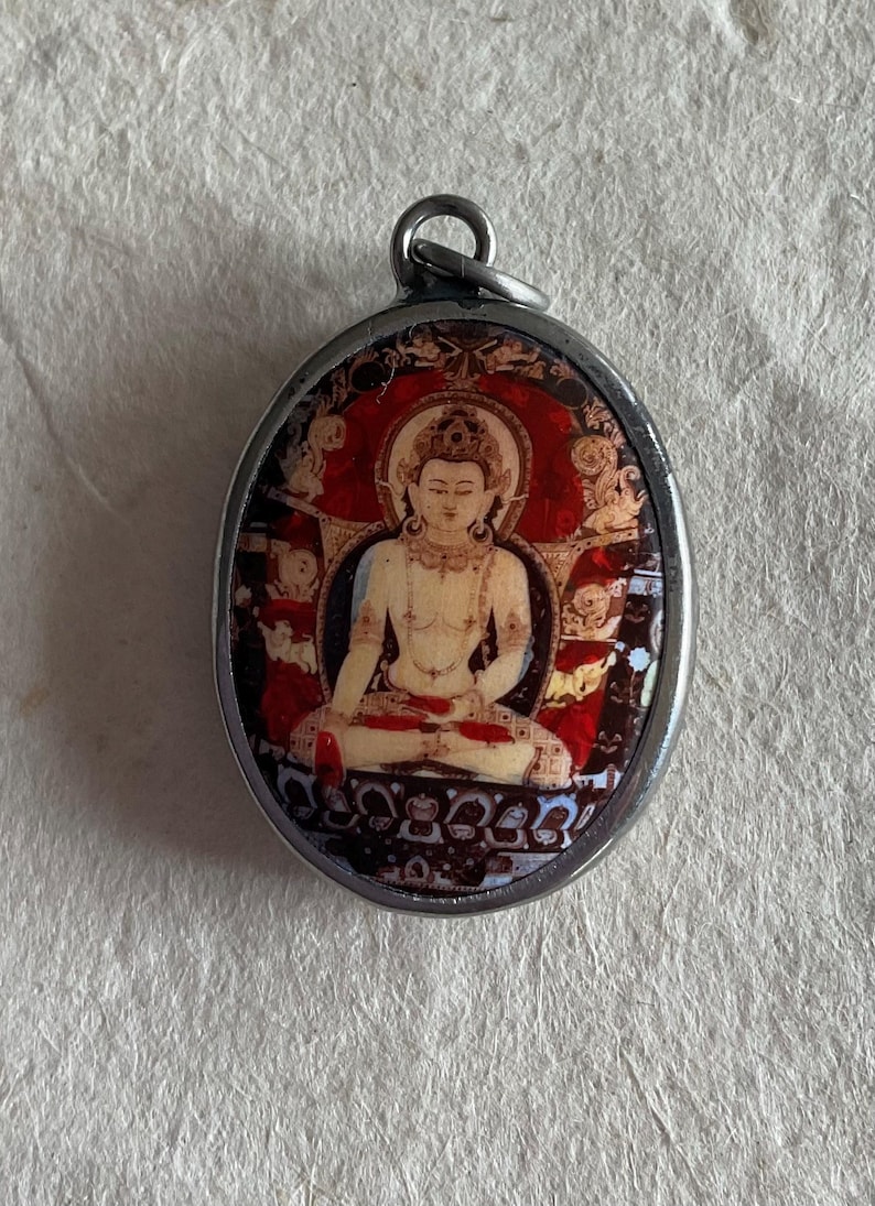 May include: A silver oval pendant with a colorful image of a seated Buddha figure. The Buddha is depicted in a red and gold robe with a halo around their head. The pendant has a detailed border with intricate designs.