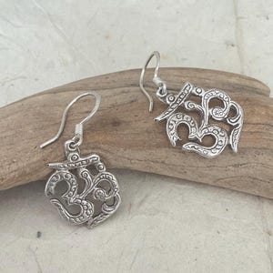 May include: A pair of silver-colored earrings featuring the Om symbol. The earrings have a detailed, openwork design with a hook closure. The earrings are displayed on a piece of driftwood against a neutral background.