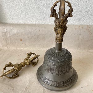 Puede incluir: Una campana antigua de bronce con intrincadas tallas y un asa detallada, junto a un cetro de mano a juego. La campana tiene una base ancha y un borde decorativo. El cetro tiene un diseño simétrico.
