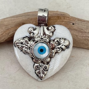 May include: Heart-shaped pendant with a white base and ornate silver-colored metalwork. The pendant features a central blue eye design and a silver bail for hanging. The pendant is approximately 2 inches in length.