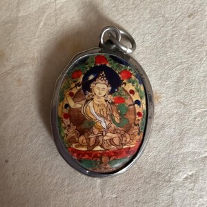 May include: A silver oval pendant with a colorful image of a deity sitting in a lotus position. The image is surrounded by a blue, red, and gold border.