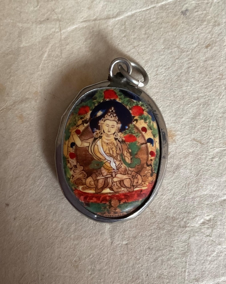 May include: A silver oval pendant with a colorful image of a deity sitting in a lotus position. The image is surrounded by a blue, red, and gold border.