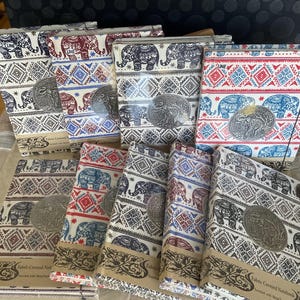 May include: A stack of fabric-covered notebooks with a repeating elephant and geometric pattern. The notebooks are decorated with a metal embellishment and the text "Fabric Covered Notebook" is printed on the bottom of each notebook.