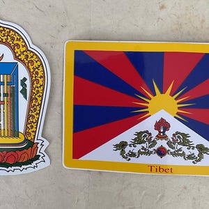 May include: Two stickers, one with a Tibetan prayer wheel and the other with the Tibetan flag. The flag is red, blue, and white with a sun and a symbol in the center. The text "Tibet" is printed below the flag.
