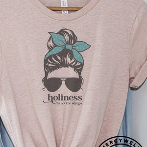 May include: A pink t-shirt with a graphic of a woman with a messy bun, sunglasses, and a teal bandana. The text "holiness is not for wimps" is printed below the graphic. The shirt is on a hanger with a blue denim shirt and a brown hat in the background.