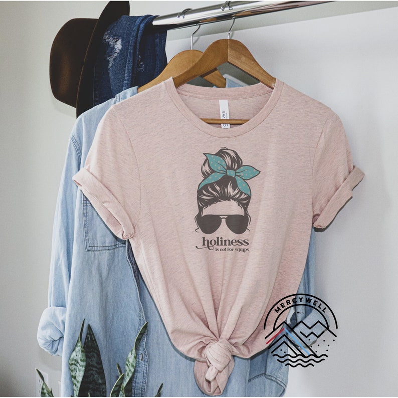 May include: A pink t-shirt with a graphic of a woman with a messy bun, sunglasses, and a teal bandana. The text "holiness is not for wimps" is printed below the graphic. The shirt is on a hanger with a blue denim shirt and a brown hat in the background.