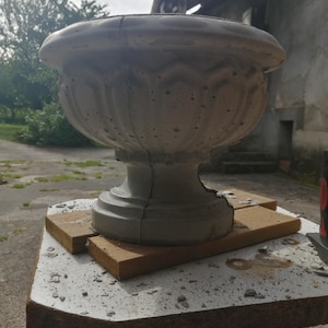 May include: A light grey concrete planter with a wide, rounded bowl shape and a decorative scalloped pattern around the exterior. The planter features a sturdy, pedestal-style base and a smooth, thick rim, showcasing a porous, textured surface.