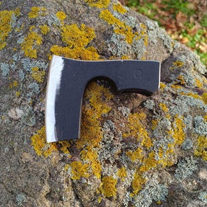 Könnte beinhalten: Eine schwarze Axt mit polierter silberner Klingenkante liegt auf einem Felsen, der mit gelbem und grauem Flechten bedeckt ist. Die Axt hat einen gebogenen Griff und eine quadratische Markierung an der Seite.
