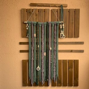Op de afbeelding: Een macramé wandkleed met groene en paarse koorden, versierd met houten kralen en decoratieve knopen. Het stuk is opgehangen aan een houten deuvel en hangt tegen een achtergrond van houten planken, wat een bohemien esthetiek creëert.