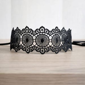 Op de afbeelding: Een zwarte kanten choker ketting met een ingewikkeld, opengewerkt ontwerp. De choker heeft een herhalend patroon van cirkelvormige motieven en delicate bloemendetails. De ketting wordt gepresenteerd op een lichtgekleurd houten oppervlak.