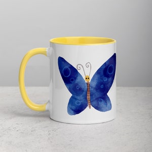 May include: A white ceramic mug with a yellow rim features a blue watercolor butterfly with brown antennae and a yellow face. The butterfly is centered on the mug.