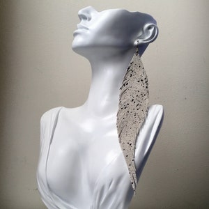 May include: A pair of long, white feather earrings with black speckles. The earrings are dangling from a mannequin's ear.