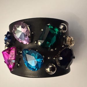 Könnte beinhalten: Schwarzes Lederarmband mit einer Vielzahl farbiger, facettierter Edelsteine. Die Edelsteine umfassen rosa, blaue, grüne, lila und klare Steine, die jeweils mit silberfarbenem Metall gefasst sind. Ein breites Statement-Armband.
