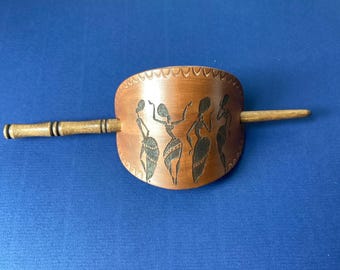 May include: A brown leather hair stick with a carved design of four women dancing. The stick is made of wood and has a natural finish.