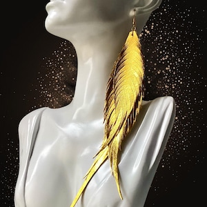 May include: A single gold feather earring is shown on a white mannequin. The earring is long and detailed, with a textured surface and a hook for the ear. The background is black with gold glitter.