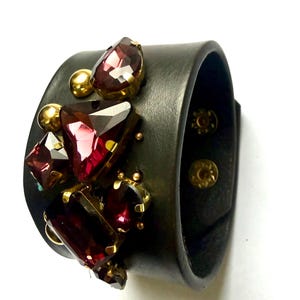Puede incluir: Pulsera de puño de cuero negro adornada con grandes piedras preciosas facetadas de color burdeos y pequeños tachuelas doradas. La pulsera tiene una banda ancha y un cierre de botón a presión. Una pieza llamativa.