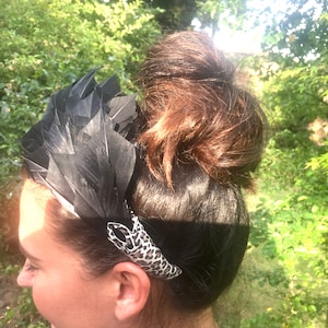 May include: A black feather headband with a leopard print accent. The headband is worn on a person with brown hair pulled back in a bun.