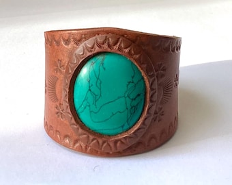 May include: A brown leather cuff bracelet with a turquoise stone inlay. The bracelet has a decorative pattern around the stone.
