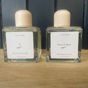 K&ouml;nnte beinhalten: Zwei durchsichtige Glasflaschen mit Holzverschl&uuml;ssen und Etiketten. Die Etiketten lauten "O & H. Candles" und die D&uuml;fte sind "Si" und "Peony & Blush". Beide Flaschen sind mit "Hand-Poured Soy Candles" gekennzeichnet.