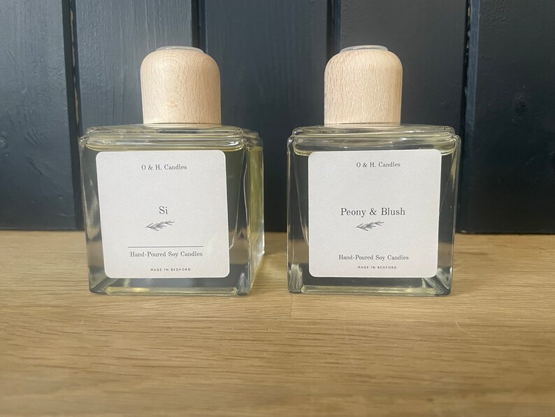 K&ouml;nnte beinhalten: Zwei durchsichtige Glasflaschen mit Holzverschl&uuml;ssen und Etiketten. Die Etiketten lauten "O & H. Candles" und die D&uuml;fte sind "Si" und "Peony & Blush". Beide Flaschen sind mit "Hand-Poured Soy Candles" gekennzeichnet.