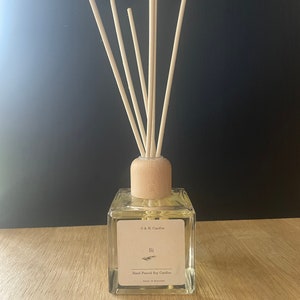 K&ouml;nnte beinhalten: Eine klare Glasdiffuserflasche mit einem Holzdeckel und nat&uuml;rlichen Schilfrohrst&auml;ben. Die Flasche hat ein Etikett mit der Aufschrift "O & E Candles" und "Si" mit einem kleinen Zweig Gr&uuml;n. Das Etikett lautet auch "Hand-Poured Soy Candles" und "Made in England".