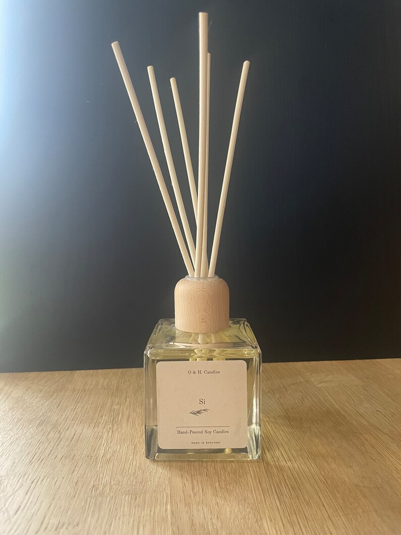 K&ouml;nnte beinhalten: Eine klare Glasdiffuserflasche mit einem Holzdeckel und nat&uuml;rlichen Schilfrohrst&auml;ben. Die Flasche hat ein Etikett mit der Aufschrift "O & E Candles" und "Si" mit einem kleinen Zweig Gr&uuml;n. Das Etikett lautet auch "Hand-Poured Soy Candles" und "Made in England".