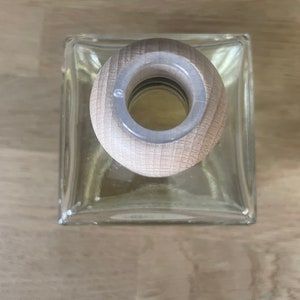 K&ouml;nnte beinhalten: Eine klare Glasflasche mit einem Holzstopfen. Die Flasche ist quadratisch und hat einen abgerundeten oberen Teil. Der Stopfen ist aus hellbraunem Holz und hat eine kreisf&ouml;rmige &Ouml;ffnung in der Mitte.