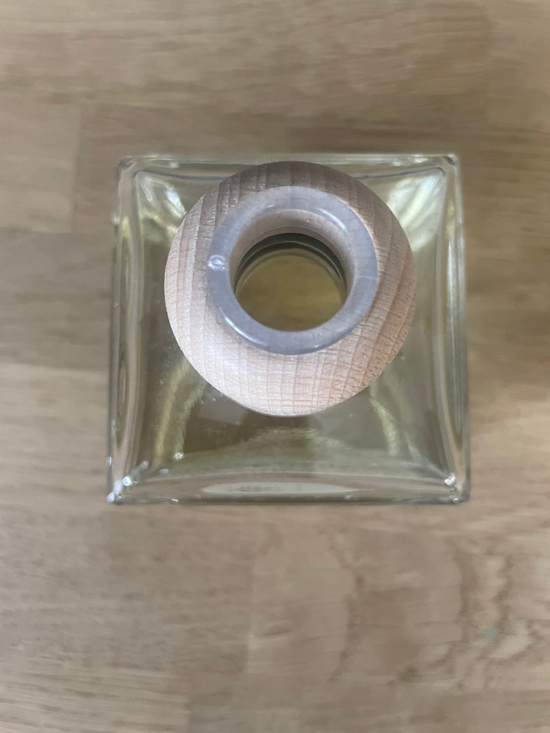 K&ouml;nnte beinhalten: Eine klare Glasflasche mit einem Holzstopfen. Die Flasche ist quadratisch und hat einen abgerundeten oberen Teil. Der Stopfen ist aus hellbraunem Holz und hat eine kreisf&ouml;rmige &Ouml;ffnung in der Mitte.