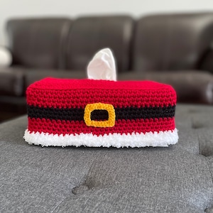 May include: A crocheted tissue box cover in the shape of a Santa Claus belt. The cover is red with a black belt and a gold buckle. The cover has a white fluffy trim around the bottom.