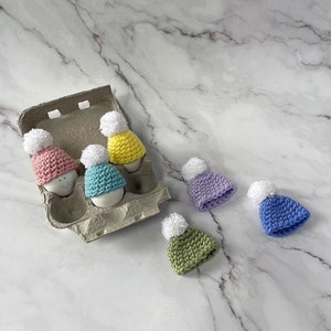 May include: Six crocheted hats in various colors, including pink, blue, yellow, and green, are displayed in an egg carton. The hats are made with a white yarn and have a pom-pom on top.