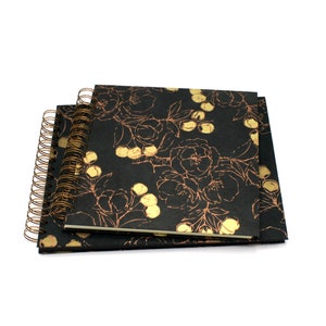 Può includere: Due quaderni quadrati con rilegatura a spirale e copertine nere caratterizzate da un motivo floreale e di bacche in oro metallizzato. I quaderni sono impilati e mostrano una rilegatura a filo color bronzo sul lato sinistro.